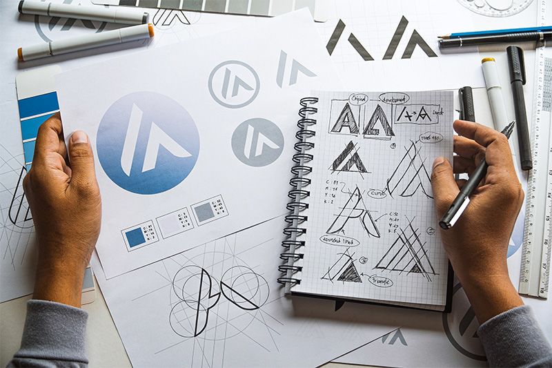 Above View of a Left Hand Holding a Sheet with Logo Concepts and a Right Hand Holding a Pen and Notebook with Sketched Logos
