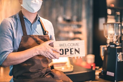 Business owner wearing a mask and holding a sign that says “Welcome – Open” to greet customers.
