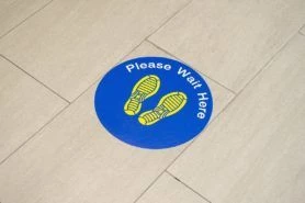 Floor sign that is a blue circle with yellow shoeprints inside that says “please wait here”