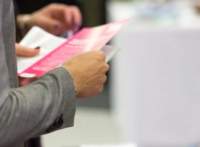 Close Up of a Womans Torso in a Gray Business Suit Holding a Pink Flyer with White Text on It