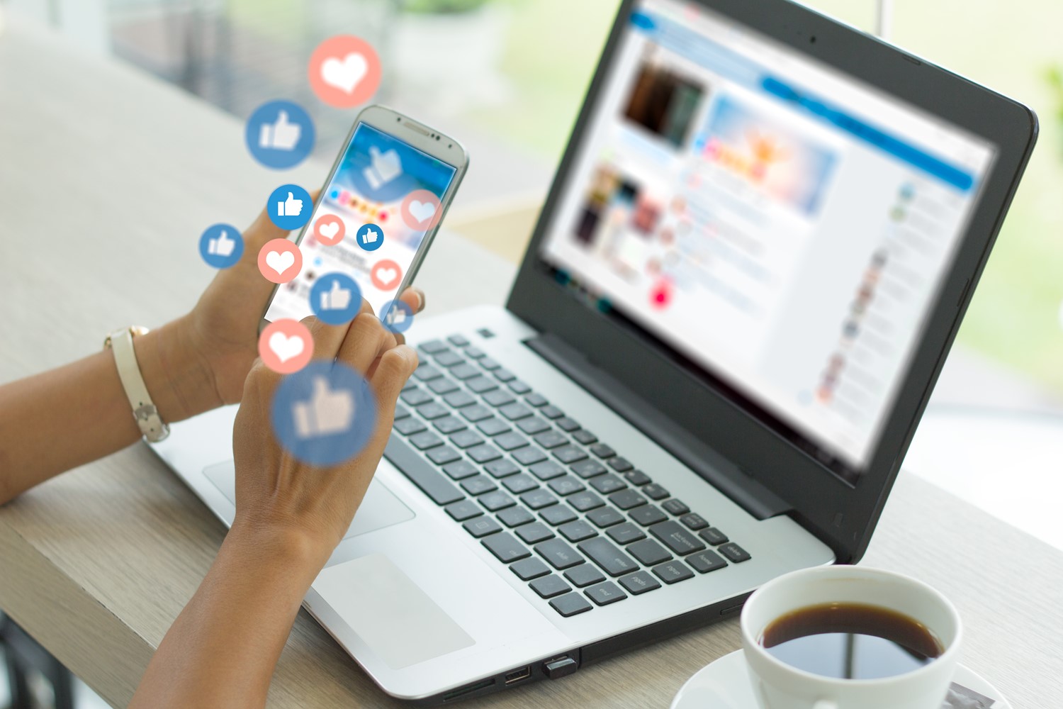 Conceptual Image of Hands Holding a Smartphone with a Laptop in Front of them and Social Media Icons Floating Above the Phone