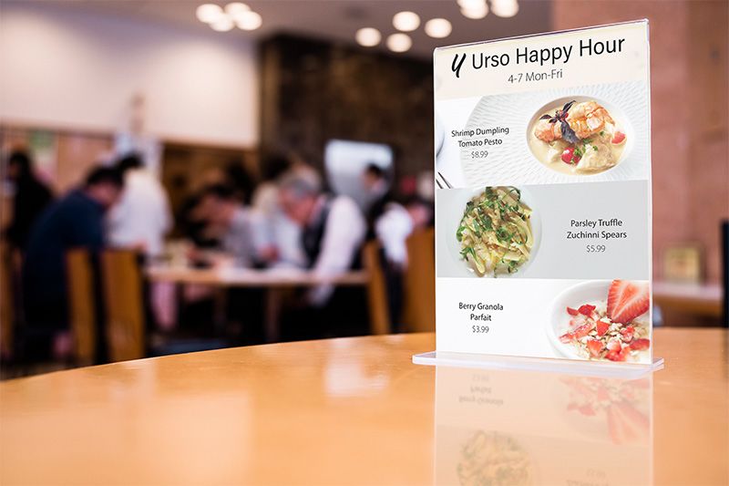 A Tabletop Sign with Happy Hour Menu Items on it on a Table with Patrons in a Restaurant Blurry in the Background
